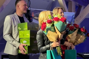 Na scenie stoją trzy osoby, dwie trzymają bukiety kwiatów i torby, a jedna niesie pudełka, w tle widać oświetlenie i sprzęt.