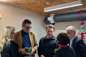 Kilka osób stoi w pomieszczeniu pod ceglanym sklepieniem i rozmawia, część trzyma kubki, w tle widoczna instalacja i dekoracje.