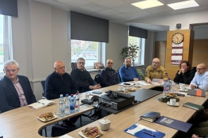 Grupa osób siedzących przy stole konferencyjnym podczas spotkania, z dokumentami, notesami i napojami, w jasnej sali z oknami i zegarem na ścianie.
