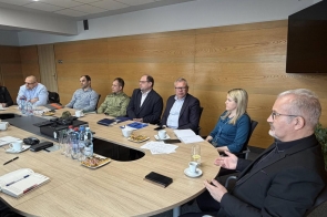 Osoby siedzące przy stole konferencyjnym podczas spotkania na tle drewnianej ściany i ekranu.