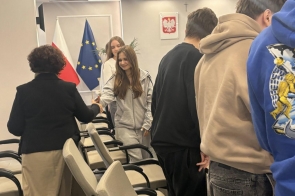 Osoby stojące w sali i witające się uściskiem dłoni, w tle rzędy krzeseł oraz flagi Polski i Unii Europejskiej.