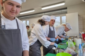 Kilka osób w strojach kucharskich przygotowuje potrawy przy stanowiskach roboczych, krojąc składniki i pracując przy blacie.