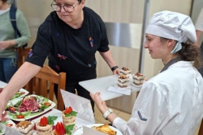 Dwie osoby stoją przy stole z przygotowanymi potrawami i deserami, układając dania na talerzach.