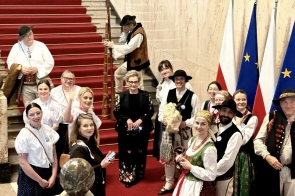 Na środku stoi Beata Macura Członkini Zarządu, a obok zgromadzona jest grupa osób, w tym część w strojach ludowych, ustawiona przed czerwonymi schodami.