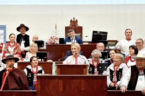  Aneta Legierska-Bujok Przewodnicząca Komisji Kultury, Sportu i Promocji Powiatu przemawia przy mównicy, wokół niej siedzą i stoją osoby, część w strojach ludowych; w tle sala obrad z drewnianymi ławami i stanowiskami.
