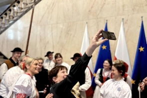Grupa osób w strojach ludowych robi wspólne zdjęcie telefonem, scena w jasnym wnętrzu z marmurową ścianą, schodami oraz flagami.
