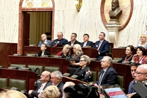 Osoby siedzące na sali obrad na podwyższonych rzędach ławek, w tle marmurowe ściany, drewniane elementy oraz rzeźba na ścianie.