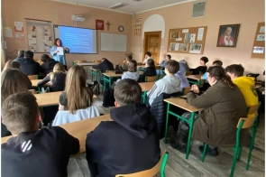 Zdjęcie przedstawia salę lekcyjną z uczniami siedzącymi w ławkach. Uczniowie zwróceni są w stronę tablicy, gdzie wyświetlana jest prezentacja. Na ścianach widoczne są plakaty edukacyjne. W klasie znajdują się ławki, krzesła i sprzęt szkolny.