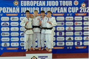 Czterech młodych mężczyzn w strojach judo stoi na podium, prezentując zdobyte medale. Jeden z nich ma złoty medal, pozostali srebrne i brązowe.