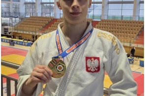 Młody mężczyzna w stroju judo stoi na trybunach hali sportowej i pokazuje medal trzymany w dłoni. W tle widoczna jest mata do walki oraz trybuny.