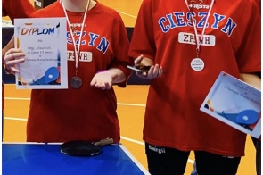 Dwie dziewczyny w czerwonych koszulkach stoją przy stole do tenisa stołowego. Mają na szyjach medale i trzymają dyplomy.