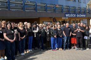 Grupa ludzi stoi przed wejściem do budynku, ustawiona wzdłuż chodnika, w słoneczny dzień.