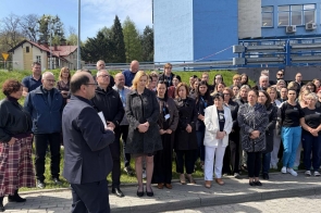 Grupa ludzi stoi przed budynkiem i słucha mężczyzny przemawiającego, przy chodniku i zielonym terenie.