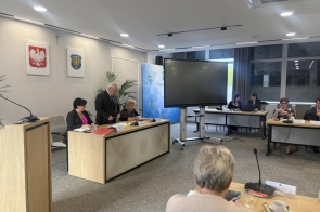 Trzy osoby przy stole, jedna osoba stoi i przemawia do mikrofonu. W tle ekran oraz flagi.