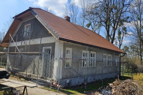 Budynek w trakcie remontu, otoczony rusztowaniami, z nowym czerwonym dachem i surową elewacją; wokół leżą materiały budowlane, a w tle widać drzewa bez liści w słoneczny dzień.