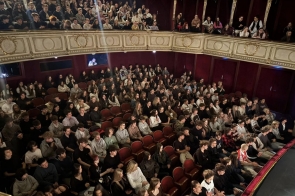 Widownia w teatrze wypełniona ludźmi siedzącymi na czerwonych fotelach, z dodatkowymi osobami na balkonie, w otoczeniu ozdobnych balustrad i czerwonych ścian.