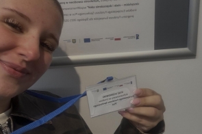 Kobieta trzyma kartkę z odręcznymi notatkami i rysunkami, uśmiecha się do aparatu. W tle tablica z zapisanymi informacjami.