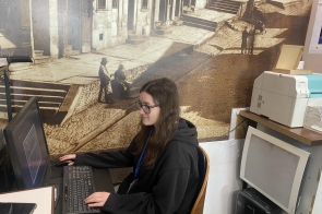 Kobieta siedzi przy biurku i pracuje na komputerze. W tle duża grafika na ścianie. Na blacie znajdują się klawiatura, mysz i inne akcesoria biurowe.