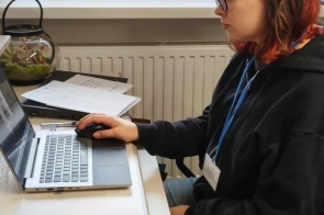Kobieta siedzi przy biurku i pracuje na komputerze. Obok znajdują się dokumenty i sprzęt biurowy. Patrzy na ekran, ręce ma na klawiaturze.