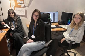 Trzy osoby siedzą w pomieszczeniu biurowym. Na pierwszym planie kobieta w ciemnej bluzie siedzi na krześle obrotowym. W tle biurka, komputery i sprzęt biurowy.