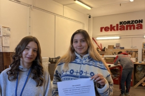 Dwie kobiety stoją w jasnym wnętrzu. Jedna trzyma kartkę z odręcznymi notatkami. W tle widoczne są osoby, stoły i materiały ustawione w sali.