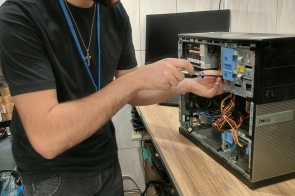 Młody mężczyzna stoi przy stanowisku roboczym i montuje elementy elektroniczne. Używa narzędzia ręcznego. Na stole widoczne są przewody i części, a w tle półki z wyposażeniem warsztatowym.