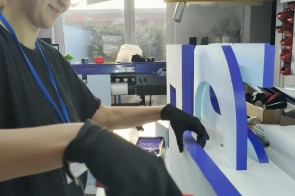 Na zdjęciu mężczyzna w okularach ochronnych i rękawicach obsługuje maszynę w hali produkcyjnej. Ustawia element przy stanowisku roboczym. W tle widoczne są inne urządzenia i wyposażenie warsztatu