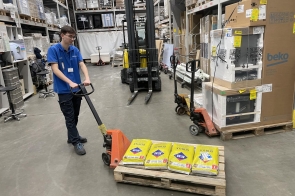 Pracownik w hali produkcyjnej pcha wózek z pojemnikami. Porusza się między stanowiskami z maszynami i urządzeniami. W tle widoczne są linie produkcyjne oraz elementy wyposażenia zakładu.