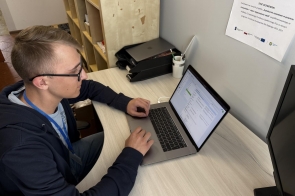 Młody mężczyzna siedzi przy biurku i pracuje przy laptopie. Obok znajduje się dodatkowy ekran. Na biurku leżą dokumenty i akcesoria, w tle widoczne jest wyposażenie miejsca pracy.