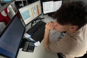 Młoda osoba siedzi przy stanowisku komputerowym i pracuje przy laptopie. Na biurku znajdują się dokumenty i akcesoria biurowe. W tle widoczne są urządzenia oraz wyposażenie miejsca pracy.