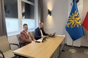 Dwóch mężczyzn siedzi przy stole podczas spotkania – jeden pracuje na laptopie, a obok stoją flagi.