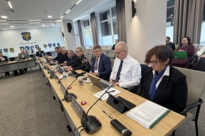Grupa osób siedzi przy długim stole podczas oficjalnego posiedzenia – mają przed sobą dokumenty, laptopy i mikrofony.