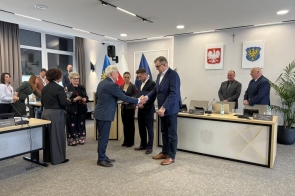 Dwóch mężczyzn w garniturach podaje sobie ręce podczas oficjalnej uroczystości, w tle stoją inni uczestnicy i widoczne są flagi oraz godła.