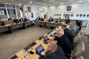 Kilka osób siedzi przy ustawionych w kształt podkowy stołach, przed nimi znajdują się tablety, dokumenty, mikrofony oraz filiżanki i butelki z wodą.