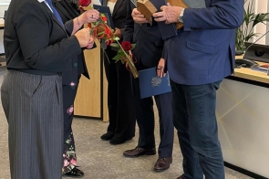 Kilka osób w eleganckich strojach uczestniczy w uroczystości wręczenia nagród – kobieta przekazuje czerwone róże, a mężczyzna trzyma pamiątkowe statuetki i dyplomy.