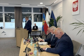 Mężczyzna stoi przy mównicy, a inni siedzą przy stole z laptopami i dokumentami; w tle widoczne flagi.