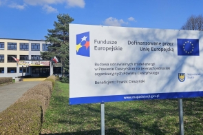Na pierwszym planie stoi tablica informacyjna o projekcie. W tle widać budynek szkoły z wejściem, flagami oraz otoczeniem z zielenią i krzewami.