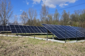 Rząd paneli fotowoltaicznych ustawionych na konstrukcjach, ogrodzony teren porośnięty trawą, w tle drzewa i linie energetyczne pod niebieskim niebem.