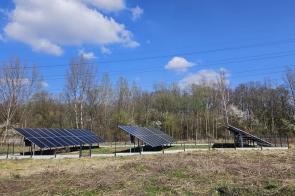 Trzy sekcje paneli fotowoltaicznych ustawionych w odstępach na ogrodzonym terenie, wokół trawa i niska roślinność, w tle pas drzew oraz linie energetyczne na tle nieba z rozproszonymi chmurami.