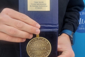 Zbliżenie na złoty medal „Olimpiada Geograficzna – finalista” trzymany w dłoniach, umieszczony w granatowym, otwartym etui.