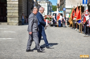 Dwóch mężczyzn w garniturach idzie brukowaną ulicą, jeden z nich niesie wiązankę kwiatów. Po prawej stronie stoi szpaler osób ze sztandarami i flagami, a w tle widoczny jest samochód.