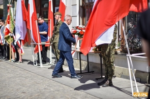 Mieczysław Szczurek Wicestarosta Cieszyński niesie wiązankę kwiatów i podchodzi do stołu ustawionego przy budynku. Obok stoją żołnierze w mundurach, a w tle widoczne są osoby trzymające flagi i sztandary.