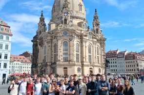 Grupa uczniów pozuje do zdjęcia na placu przed barokowym kościołem. W tle widoczna zabytkowa zabudowa oraz turyści spacerujący po rynku.