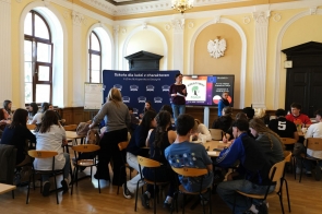 Uczniowie siedzą przy stolikach i piszą na kartkach, a osoba stojąca z przodu mówi do grupy, obok widoczny ekran z wyświetloną prezentacją.