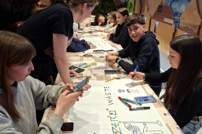 Uczniowie siedzą przy stole i wspólnie tworzą plakat o tematyce zero waste, korzystając z markerów i telefonów, podczas zajęć warsztatowych w sali edukacyjnej.
