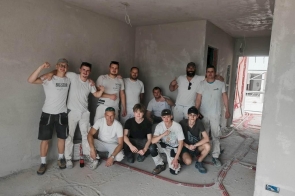 Grupa mężczyzn i chłopaków stoi w niewykończonym wnętrzu budynku, pozując do zdjęcia wśród surowych ścian i instalacji na podłodze.