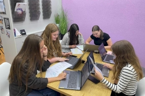Grupa uczennic siedzi przy stole i pracuje na laptopach oraz kartkach, przeglądając materiały i notatki.