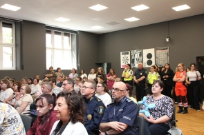 Na sali siedzą dzieci, młodzież i dorośli. Wśród uczestników znajdują się funkcjonariusze policji, straży miejskiej oraz ratownicy medyczni, a część osób stoi pod ścianą i obserwuje wydarzenie.