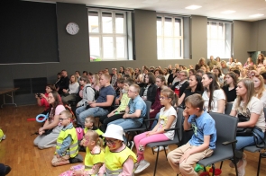 Na sali siedzą dzieci, młodzież i dorośli obserwujący występ. W pierwszych rzędach znajdują się dzieci w kamizelkach odblaskowych, a część osób robi zdjęcia telefonami.