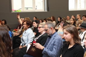 Na sali siedzą młodzi ludzie i osoby dorosłe. Część uczestników trzyma dokumenty i teczki, a jedna z osób robi zdjęcie telefonem.